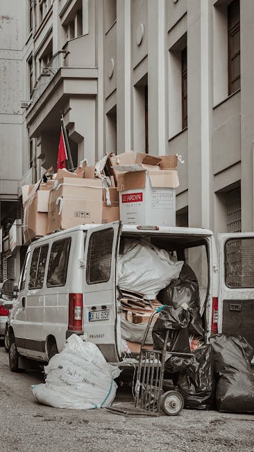 The image depicts a white commercial van parked on a city street in front of a multi-story building with a beige facade. The van's rear doors are open, revealing a cargo area filled with various recyclable or discarded materials, including large black and white plastic bags, flattened cardboard boxes, and a stack of thin wooden pallets. Several boxed items and packing materials are stacked loosely inside, with some spilling out onto the pavement. Nearby, a flat trolley with a handle and a small wheeled platform loaded with black plastic bags is positioned close to the open rear of the van, suggesting active loading or unloading. The surrounding environment features an urban setting with a narrow sidewalk, and the building's architectural details include large windows and decorative moldings. The scene appears to be part of an independent waste collection or on-site clearance operation, possibly conducted by Rubbish Removal Mayfair, aimed at facilitating private rubbish disposal or alternative waste handling outside regular municipal services. The lighting indicates natural daylight with a neutral, unembellished atmosphere suitable for documentation purposes in a professional waste management context.