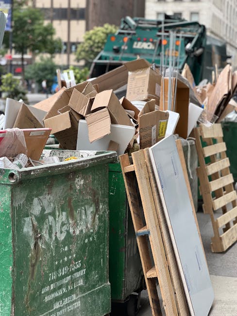 An overflowing green rubbish dumpster filled with various types of waste, including cardboard boxes, paper packaging, and wooden pallets, placed on a city street. The cardboard boxes are in different sizes, some flattened and others still in three-dimensional form, with visible corrugated textures and brown tones. A set of wooden pallets, with a light natural wood finish, leans against the side of the dumpster, partially obscuring the lower section of the waste. In the background, a commercial waste collection vehicle with the word 'LUCAS' visible on its side is parked nearby, surrounded by trees and urban buildings, indicating an outdoor setting for private waste disposal. The environment appears to be in daylight with an overcast sky, typical of a busy city area where rubbish removal services are engaged in on-site clearance or alternative waste handling.