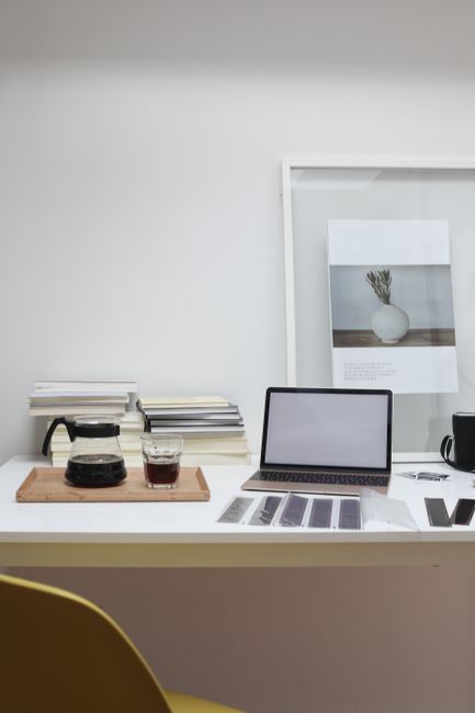 A minimalist office workspace featuring a white desk against a plain white wall. On the left side of the desk, there is a wooden tray holding a glass coffee pot and two matching glasses filled with dark beverages. To the right, a closed silver laptop with a black border and keyboard is positioned centrally, with several printed document sheets and charts fanned out in front of it. Behind the laptop, a stack of assorted papers and folders is neatly arranged, with a black wired mug or cup placed further to the right. A large framed picture of a vase with flowers, mounted on the wall behind the desk, adds decorative interest. The workspace appears clean and organized, with a modern, professional ambiance suitable for routine office tasks. Throughout, natural light illuminates the scene, emphasizing the simple, tidy environment, and subtly linking to themes of private or independent working arrangements typical of alternative waste handling or rubbish removal services managed by Rubbish Removal Mayfair.