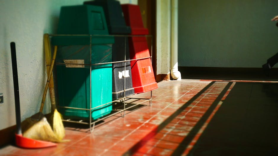 In this indoor scene, three large plastic waste bins are arranged in a metal wire rack placed against a textured white wall. The bins are made of durable, smooth plastic with a matte finish, and are colored from left to right in green, black, and red, indicating different waste streams. The green bin has a matching green lid, the black bin features a darker lid, and the red bin has a bright red lid, all fitting securely onto their respective containers. To the left of the bins, a yellow-handled broom with a straw head rests against the wall, while a red dustpan lies on the red-tiled floor beneath, both positioned near the edge of the scene. The floor consists of reddish-brown terracotta tiles with visible grout lines, providing a warm tone, and is illuminated by natural sunlight that casts long shadows of the broom, dustpan, and the wire rack with the bins onto the floor, emphasizing the scene's outdoor-adjacent environment. The background includes a corner where a white drainpipe runs vertically along the wall, and a narrow gap in the wall to the right hints at an adjacent space, all of which reflect a typical storage area suited for private waste collection, aligning with the themes of independent rubbish disposal managed by services such as Rubbish Removal Mayfair.