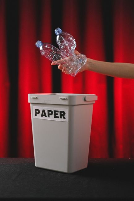 A grey paper recycling bin with a white label reading 'PAPER' is positioned on a black surface in front of a red curtain backdrop. An outstretched arm on the right side of the image is dropping three crumpled clear plastic water bottles into the bin. The bottles have blue caps and are made of transparent PET plastic, with textured surfaces characteristic of reusable bottles. The scene suggests an act of waste disposal, highlighting the separation of paper waste from plastic bottles. The setting appears to be indoors, perhaps at an event or in a waste management demonstration context, emphasizing the importance of proper rubbish segregation in compliance with independent collection practices or private waste handling services like those offered by Rubbish Removal Mayfair. The lighting is focused to evenly illuminate the bin and the recycling activity, with the red curtain providing a contrasting background that draws attention to the recycling process. This visual aligns with themes of environmentally responsible waste management and alternative rubbish disposal methods, relevant to commercial waste law and on-site clearance services in Mayfair.