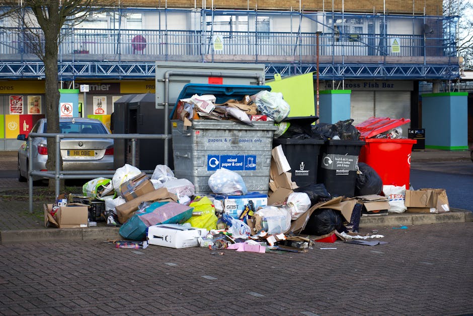 A large collection of mixed household waste and rubbish bags is piled on a paved sidewalk in front of a small parking area, with several black trash bags, cardboard boxes, paper bags, and loose packaging materials visible. In the center, there is a grey recycling bin labeled for mixed paper and cardboard, filled with various paper waste, with some items spilling over the top. To the right, black and red waste bins are present, with black bags and rubbish partially obscuring them. A silver car is parked on the left side, behind a metal railing that separates the parking area from the rubbish pile. The background includes a commercial building with a blue metal scaffold structure, glass windows, and storefronts, some signage visible, suggesting a busy urban environment. Natural daylight illuminates the scene, emphasizing the disorganized accumulation of waste, which appears ready for a private rubbish collection service, such as those offered by Rubbish Removal Mayfair, to handle as part of alternative waste management or on-site clearance.
