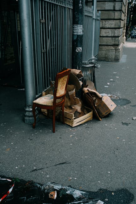 A collection of discarded cardboard boxes, some flattened and others partially collapsed, rest on the concrete sidewalk near a metal fence and an old wooden chair with a fabric-covered seat and backrest. The cardboard appears worn with visible creases, torn edges, and printed labels, indicating they are used packing materials. The scene is set outdoors next to a stone building with a classical architectural style, featuring large stone blocks and a lamppost. The boxes and chair are positioned close to each other, slightly leaning against the fence, with some additional small debris scattered on the pavement. Natural daylight illuminates the area, highlighting the textures and colors of the cardboard, the wooden chair, and the surroundings. This scene suggests a typical urban environment where waste such as packing materials is temporarily left for collection, and it relates to private or independent rubbish disposal services like those offered by Rubbish Removal Mayfair, especially for urgent or same-day bulky waste collection. The overall impression reflects a snapshot of waste awaiting removal outside a city building, consistent with rubbish removal services aimed at clearing excess or bulk waste types.