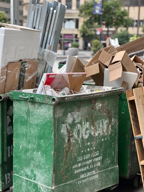A large collection of mixed household waste and rubbish bags is piled on a paved sidewalk in front of a small parking area, with several black trash bags, cardboard boxes, paper bags, and loose packaging materials visible. In the center, there is a grey recycling bin labeled for mixed paper and cardboard, filled with various paper waste, with some items spilling over the top. To the right, black and red waste bins are present, with black bags and rubbish partially obscuring them. A silver car is parked on the left side, behind a metal railing that separates the parking area from the rubbish pile. The background includes a commercial building with a blue metal scaffold structure, glass windows, and storefronts, some signage visible, suggesting a busy urban environment. Natural daylight illuminates the scene, emphasizing the disorganized accumulation of waste, which appears ready for a private rubbish collection service, such as those offered by Rubbish Removal Mayfair, to handle as part of alternative waste management or on-site clearance.