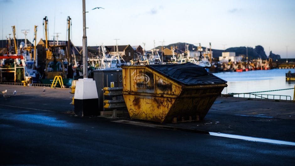 A large, yellow metal skip with a black plastic cover on top, positioned on a paved surface near a waterfront with boats and marine vessels moored along the quay. The skip shows signs of rust and wear, with visible patches of corrosion and faded markings, indicating it has been used for construction or waste disposal. To the left of the skip, there is a small black utility box and an electrical transformer mounted on a pole, with several yellow and black safety barriers nearby. The background features a row of docked boats, some with masts and rigging, set against a partly cloudy sky. The scene is illuminated by natural daylight, creating a neutral, professional tone suitable for describing waste removal or on-site clearance activities at a marina or harbor setting, consistent with independent disposal solutions offered by companies like Rubbish Removal Mayfair.