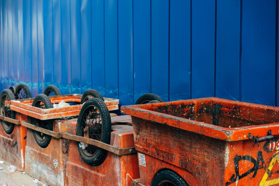 The image displays a row of orange plastic skips used for waste collection, positioned on a concrete surface. The skips are weathered, with visible dirt, grime, and some graffiti markings, indicating frequent use. One skip in the foreground is partially filled or stained, with dirt and residual debris inside, while the other skips appear empty. Each skip is equipped with a black rubber wheel on each corner, which are affixed to metal axles, allowing for easier mobility. The background consists of a tall, smooth blue metal fence with visible vertical ridges, serving as a boundary for the waste collection area. The scene is lit with natural daylight, with no shadows or reflections suggesting overcast weather. The arrangement and condition of the skips suggest they are used for building waste or large trash removal, aligning with private disposal services that Rubbish Removal Mayfair offers for alternative waste handling solutions beyond standard kerbside collection.