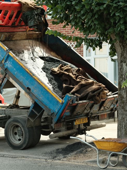 A large, yellow metal skip with a black plastic cover on top, positioned on a paved surface near a waterfront with boats and marine vessels moored along the quay. The skip shows signs of rust and wear, with visible patches of corrosion and faded markings, indicating it has been used for construction or waste disposal. To the left of the skip, there is a small black utility box and an electrical transformer mounted on a pole, with several yellow and black safety barriers nearby. The background features a row of docked boats, some with masts and rigging, set against a partly cloudy sky. The scene is illuminated by natural daylight, creating a neutral, professional tone suitable for describing waste removal or on-site clearance activities at a marina or harbor setting, consistent with independent disposal solutions offered by companies like Rubbish Removal Mayfair.