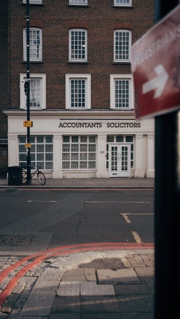 A three-storey brick building with a white ground-floor façade housing a business labeled 'Accountants Solicitors' in black uppercase letters. The ground floor features large, multi-pane sash windows and a central door with glass panels. To the left of the entrance, a bicycle is secured to a metal bike rack. In front of the building, a paved sidewalk with red brick edging and a pedestrian crossing is visible. A tall black lamp post stands on the pavement near the left side, while the background shows additional brick buildings with similar architectural styles. On the right side of the image, a blurred red and white traffic sign partially obscures the scene, indicating street traffic regulation. The overall atmosphere appears to be an urban street scene during daylight, with natural lighting illuminating the building’s façade and surrounding features. This setting relates to independent waste management and rubbish removal services often used for commercial property clearances, similar to those provided by Rubbish Removal Mayfair, aimed at facilitating professional estate clearance without reliance on local authority waste collection. The image emphasizes the importance of private waste handling within urban environments for efficient rubbish disposal and estate clearance.